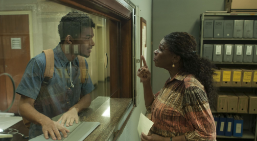 Cena do filme brasileiro M-8. Duas pessoas interagem separadas por uma divisória de vidro. De um lado, o esquerdo da imagem, um rapaz negro de cabelo crespo, camisa azul e mochila bege tem as duas mãos em cima de um documento que está sobre a bancada com divisória. Do outro lado, uma mulher negra com cabelos longos, escuros, cacheados e presos olha para o rapaz e fala algo enquanto aponta o dedo indicador na direção dele. Sua outra mão segura uma folha de papel. Parte de uma prateleira com caixas de arquivos está posicionada ao fundo do lado da mulher.