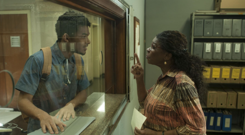 Cena do filme brasileiro M-8. Duas pessoas interagem separadas por uma divisória de vidro. De um lado, o esquerdo da imagem, um rapaz negro de cabelo crespo, camisa azul e mochila bege tem as duas mãos em cima de um documento que está sobre a bancada com divisória. Do outro lado, uma mulher negra com cabelos longos, escuros, cacheados e presos olha para o rapaz e fala algo enquanto aponta o dedo indicador na direção dele. Sua outra mão segura uma folha de papel. Parte de uma prateleira com caixas de arquivos está posicionada ao fundo do lado da mulher. 