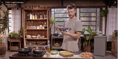 Foto de um homem segurando um prato e um garfo. Ele é branco, tem cabelos curtos loiros e veste uma camiseta bege. Ele olha para o prato em sua mão, que é azul. Na bancada à frente dele, estão duas panelas no fogão e algumas travessas de vidro com comida. Ao fundo, há um armário de madeira com utensílios e mantimentos, diversos vasos de planta e três janelas grandes.