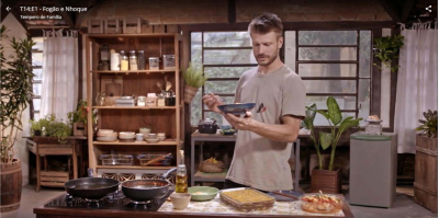 Foto de um homem segurando um prato e um garfo. Ele é branco, tem cabelos curtos loiros e veste uma camiseta bege. Ele olha para o prato em sua mão, que é azul. Na bancada à frente dele, estão duas panelas no fogão e algumas travessas de vidro com comida. Ao fundo, há um armário de madeira com utensílios e mantimentos, diversos vasos de planta e três janelas grandes. 