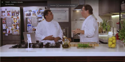 Foto de dois homens conversando na cozinha. À esquerda, está Batista, que é negro, tem cabelos curtos escuros e olha para Claude, à direita da imagem. Claude, que é branco, tem cabelos curtos escuros e usa óculos, está gesticulando e olhando para o colega. Ambos utilizam o uniforme de chefs de cozinha. Na bancada, que está ao redor deles, e ao fundo, é possível ver diversos equipamentos e utensílios da cozinha profissional.