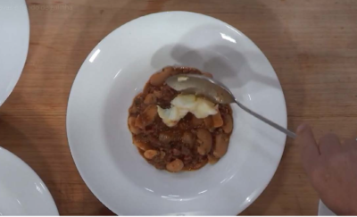 Foto de uma pessoa colocando comida em um prato. A imagem é vista de cima, é possível ver apenas uma mão branca segurando uma colher, que contém um tipo de purê. No prato, já estão alguns alimentos. O prato é redondo, fundo e branco. Ao redor dele, é possível ver a borda de outros pratos.