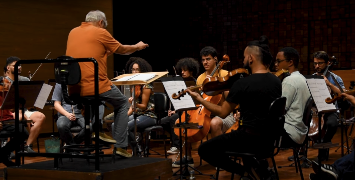 Foto de ensaio da Ocam. À esquerda, há um homem branco, de cabelos curtos e grisalhos, camiseta laranja, calça cinza e tênis marrom. Ele está sentado, regendo a orquestra. À sua frente há oito músicos tocando instrumentos como violoncelo e violino, com estantes de partituras à sua frente.