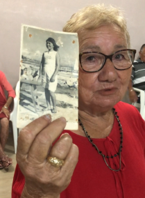 Foto de uma mulher branca idosa, de cabelos curtos, lisos e loiros claros, segurando uma foto em preto e branco de uma mulher de cabelos escuros e vestido branco na praia. A senhora usa óculos, colares pretos e uma blusa vermelha. Na mão que segura a foto, um anel dourado.