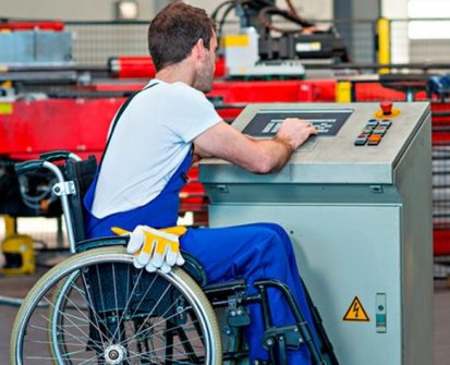 Foto de um homem em uma cadeira de rodas operando o painel de uma máquina industrial. Ele é branco, tem cabelos castanhos curtos e está com o rosto voltado para o painel. Ele está usando um macacão azul e camiseta branca, com luvas de trabalho amarelas penduradas na lateral da cadeira. O painel que ele está manuseando é metálico, com botões e uma tela na parte superior. Ao fundo, há máquinas industriais em um ambiente de fábrica, com cores predominantes em vermelho, preto e cinza.