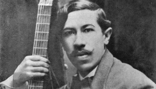 foto em preto e branco, em plano próximo do rosto, de um homem pardo de cabelos ondulados curtos e bigode. Ele segura um braço de violão.