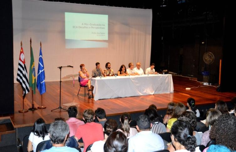 Foto de auditório com dezenas de pessoas voltadas para um palco com sete pessoas compondo uma mesa. São duas mulheres e cinco homens, em sua maioria brancos. O palco é de madeira e ao lado da mesa há três bandeiras: do Estado de São Paulo, do Brasil e da USP. No fundo há uma projeção de slide.