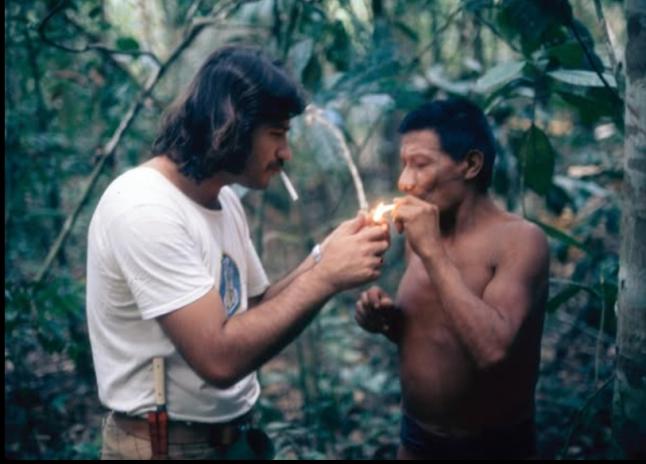 Foto de homem branco de cabelos lisos e castanhos, até os ombros, e de homem indígena de cabelos curtos e escuros fumando. O homem branco está de camiseta branca e acende o cigarro do indígena, que está sem camisa. Ao fundo, as árvores da floresta.