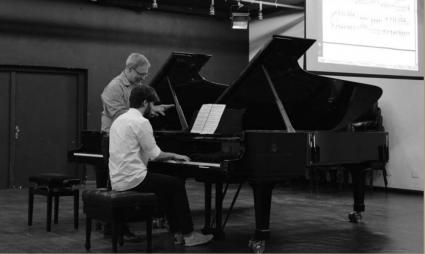 Foto em preto e branco de um homem, aparentemente jovem, de cabelos escuros e camisa branca sentado ao piano, com um professor mais velho, de cabelos grisalhos e camisa clara ao lado orientando o pianista. Ao fundo há parte de uma porta dupla e ao fundo acima há a projeção de uma partitura.