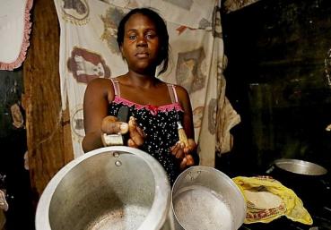 Foto de uma mulher jovem e negra que segura duas panelas vazias na direção da câmera. Ela veste uma blusinha preta estampada e olha com expressão séria para a câmera.  Atrás dela há um varal com panos e lençois pendurados.