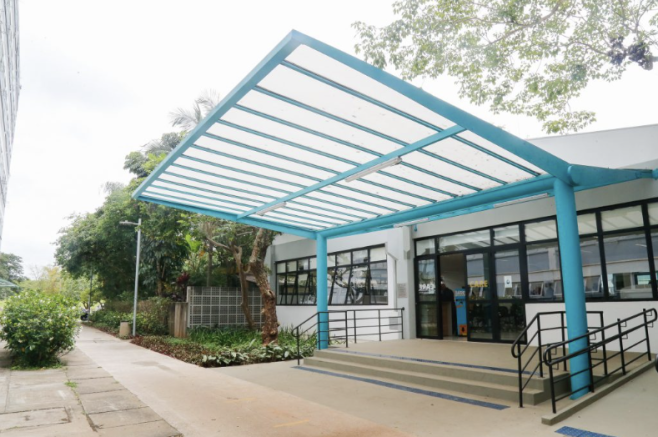Foto da fachada de um prédio branco térreo, com escada de dois degraus e rampa de acesso. A entrada do prédio possui uma cobertura com estrutura azul. Acima do telhado há galhos de uma árvore. Na frente do prédio há uma calçada com um caminho com árvores e arbustos.