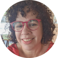 Foto de uma mulher branca, com cabelos de tamanho médio, cacheados e castanhos. Ela usa oculos de haste vermelha e blusa vermelha.