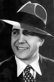 Foto em preto e branco do rosto de um homem sorrindo. Ele é branco, tem olhos escuros, veste um terno escuro, camisa branca e gravata estampada. Ele olha para o lado e usa um chapéu.