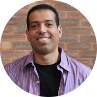 Foto de homem negro, de olhos castanhos, cabelos curtos e escuros, sorrindo. Ele veste camisa lilás com listras brancas aberta sobre uma camiseta preta. Ao fundo, uma parede de tijolos.