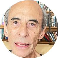 Foto de homem branco, careca, com cabelos curtos lisos e grisalhos e olhos castanhos. Ele olha para a câmera e tem a boca entreaberta. No fundo, há uma estante com livros. 