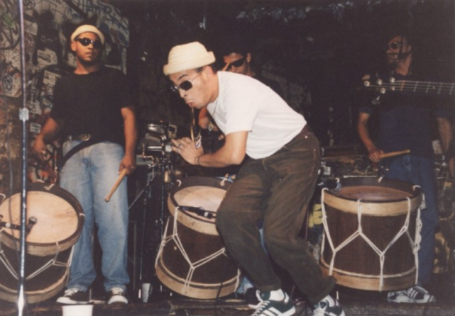 Foto de quatro homens se apresentando em um palco. O homem em primeiro plano está levemente abaixado e em movimento. Ele é branco, usa óculos escuros, touca de palha clara, camisa branca e calças pretas. Atrás dele, três homens tocam grandes tambores.