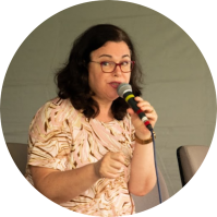 Foto de mulher branca falando ao microfone. Ela tem cabelos escuros e ondulados  na altura dos ombros, olhos castanhos, usa óculos de armação vermelha e veste um vestido estampado de ondas nas cores rosa, marrom e bege. Ao fundo, um tecido cinza.