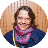 Foto de mulher branca, sorrindo. Ela tem cabelos de tamanho médio, castanhos e ondulados, olhos castanhos e usa óculos de armação transparente. Veste casaco azul e cachecol estampado nas cores rosa e azul. Atrás dela há uma parede de madeira clara.