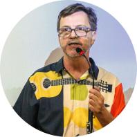 Foto de um homem branco. Ele tem cabelos curtos, lisos e castanhos, olhos castanhos, barba e bigode curtos e grisalhos e usa óculos com armação quadrada e preta. Ele fala ao microfone e veste camisa estampada com um violão e formas geométricas nas cores preto, branco, amarelo, verde e vermelho.  Atrás dele, uma tela de projeção.