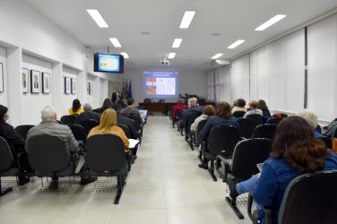 Foto de sala, tirada do fundo. A sala é dividida em dois conjuntos de cadeiras, na cor verde, posicionadas lado a lado e uma em várias fileiras. Há um corredor entre elas. No fundo da imagem, uma mesa grande de madeira, um pulpito de acrilico, de onde fala uma pessoa, e uma tela de computador projetada na parede. Um monitor de TV está posicionado no lado superior esquerdo da sala. As paredes são brancas, com quadros na parede de um lado, e persianas também brancas do outro lado. 