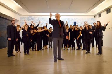 Foto dos integrantes do Coro de Câmara Comunicantus. Eles estão em uma sala. À frente do grupo, está regente e professor do coro. Ele é um homem mais velho, com cabelos curtos e brancos nas laterais da cabeça, usa um terno preto com gravata vermelha e está com uma das mãos levantadas. Ao fundo, está o grupo. Todos usam roupas pretas e olham para a câmera, com os braços levantados.