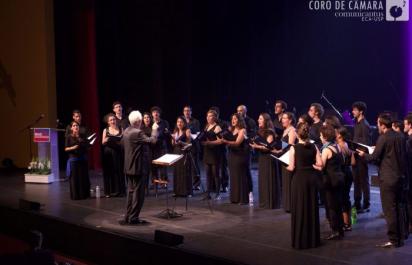 Foto de uma apresentação do Coro de Câmara da ECA. Há diversos coristas de pé em cima de um palco, usam roupas pretas e estão posicionados numa formação de meia lua, segurando partituras musicais. À sua frente há um regente, branco, de cabelos curtos e brancos, vestindo um terno preto.