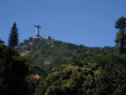 Foto da estátua do Cristo Redentor no alto do morro do Corcovado. Na parte de baixo da imagem, o telhado de uma casa rodeado de árvores. Ao fundo, um céu limpo e azul. 