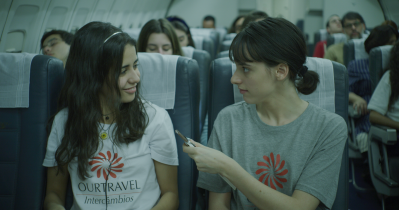 Duas garotas estão num avião. Ambas vestem camisas de intercâmbio (uma branca, outra cinza). A garota, de cabelo liso e pele branca, à esquerda oferece um chocolate a outra, morena, que levemente sorri, como quem agradece o oferecimento.