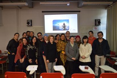 Foto de diversas pessoas em pé, posando para foto. Das dezessete pessoas, doze são mulheres. Todos vestem roupas de frio. As pessoas estão numa sala de aula e, ao fundo, está uma projeção em um telão com a frase “Mindfulness/Atenção Plena” e a foto de uma pessoa meditando.