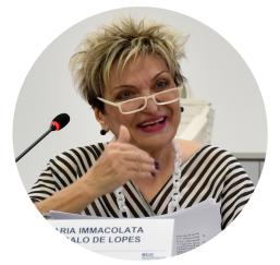 Foto da professora Maria Immacolata. Ela é uma mulher branca, com cabelo loiro, liso e curto. Usa óculos de armação retangular na cor creme, colar branco e blusa com listras pretas e brancas. Na imagem, a professora está sorrindo durante uma apresentação, e um microfone aparece no canto inferior esquerdo.