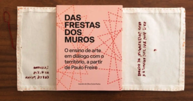  Foto da tese de doutorado encadernada como um livro. A foto exibe a capa da tese com o título, subtítulo e nome do autor em letras pretas. O fundo é rosa claro com linhas pontilhadas em um tom de rosa mais escuro, formando figuras geométricas sobrepostas. O livro está sobre o verso de um tecido branco na horizontal com frases bordadas em vermelho. Atrás desse tecido há uma superfície de madeira. 