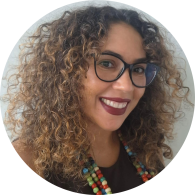 Foto de mulher negra em semi perfil sorrindo. Ela tem olhos castanhos, cabelos cacheados, longos e castanhos com as pontas claras e usa óculos com armação preta e arredondada, blusa preta, colar de bolinhas coloridas e batom vinho. O fundo é cinza. 