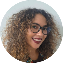 Foto de mulher negra em semi perfil sorrindo. Ela tem olhos castanhos, cabelos cacheados, longos e castanhos com as pontas claras e usa óculos com armação preta e arredondada e batom vinho. O fundo é cinza.