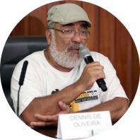 Foto de homem negro falando ao microfone. Ele usa óculos de armação quadrada, boina cinza e camisa branca. O fundo é escuro. 