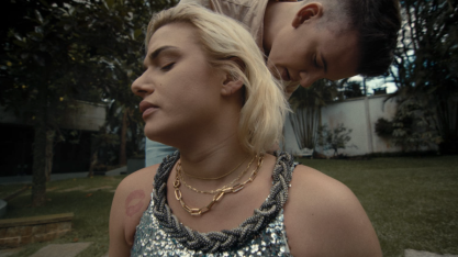 Cena do filme Dentro. Perfil do rosto de uma mulher, branca, de cabelos loiros curtos, que usa colares dourados e blusa com lantejoulas cinzas e tem uma tatuagem de beijo no ombro. Atrás dela, um homem branco de cabelo castanho e curto está em pé e inclinado em direção a mulher, que está sentada. Ele toca as costas dela. Algumas árvores formam o plano de fundo.