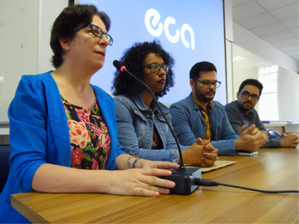 Foto de quatro pessoas sentadas à mesa de madeira castanha de um auditório, onde há uma tela com projeção ao fundo. Da esquerda para a direita: mulher branca de cabelos curtos, óculos, blusa azul e camisa florida, mulher negra de cabelos escuros crespos médios, olhos escuros, jaqueta jeans falando ao microfone, homem branco de cabelos e barba curtas e escuras e jaqueta jeans ajeitando os óculos. homem branco de cabelos e barba escuros e curtos, óculos e camisa cinza.