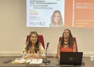foto de um seminário. Duas mulheres estão sentadas em uma mesa grande. à esquerda, uma mulher branca, de cabelos longos e castanho claros. Ela tem um óculos apoiado sobre a testa. Veste uma blusa estampada e tem os antebraços apoiado um sobre o outro. à direita, uma mulher branca, de cabelos longos e castanho claros. Ela veste uma blusa laranja e um colar de madeira, marrom. à frente dela, um notebook aberto. Ao fundo, é projetada uma imagem da palestrante, título da palestra e outras informações.