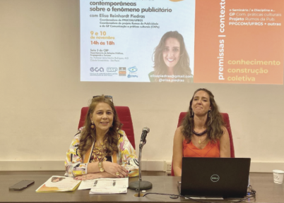 foto de um seminário. Duas mulheres estão sentadas em uma mesa grande.  à esquerda, uma mulher branca, de cabelos longos e castanho claros. Ela tem um óculos apoiado sobre a testa. Veste uma blusa estampada e tem os antebraços apoiado um sobre o outro. à direita, uma mulher branca, de cabelos longos e castanho claros. Ela veste uma blusa laranja e um colar de madeira, marrom. à frente dela, um notebook aberto. Ao fundo, é projetada uma imagem da palestrante, título da palestra e outras informações.