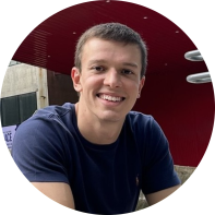 Foto de homem branco sorrindo. Ele tem olhos castanhos, cabelos curtos, lisos e castanhos e usa uma camiseta azul escura. O fundo é vermelho escuro com parte de um prédio cinza aparecendo. 