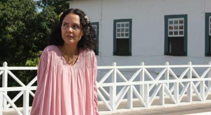 Imagem retirada de um documentário sobre Cora Coralina. Em primeiro plano, há uma mulher branca, de cabelos escuros, curtos e enfeitados. Ela usa uma roupa rosa. Ao fundo, é possível ver uma casa branca com janelas verdes e a copa de algumas árvores.