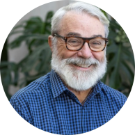 Foto de um homem branco sorrindo. Ele tem cabelos lisos, curtos e brancos, barba branca, está com óculos de armação marrom e usa uma camisa xadrez azul. Há plantas ao fundo.