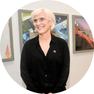 Foto de uma mulher de pele branca e cabelos curtos e brancos, usando óculos de armação redonda e uma camisa preta. Ela está sorrindo e usando um broche em formato de estrela na altura do peito. Ao fundo, há obras de arte emolduradas na parede com elementos coloridos.