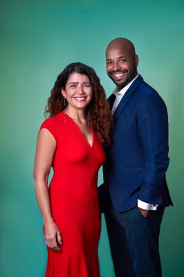 À esquerda, uma mulher branca de cabelo cacheado sorrindo, ao lado de um homem negro, careca, sorrindo e de terno azul. O fundo da foto é verde