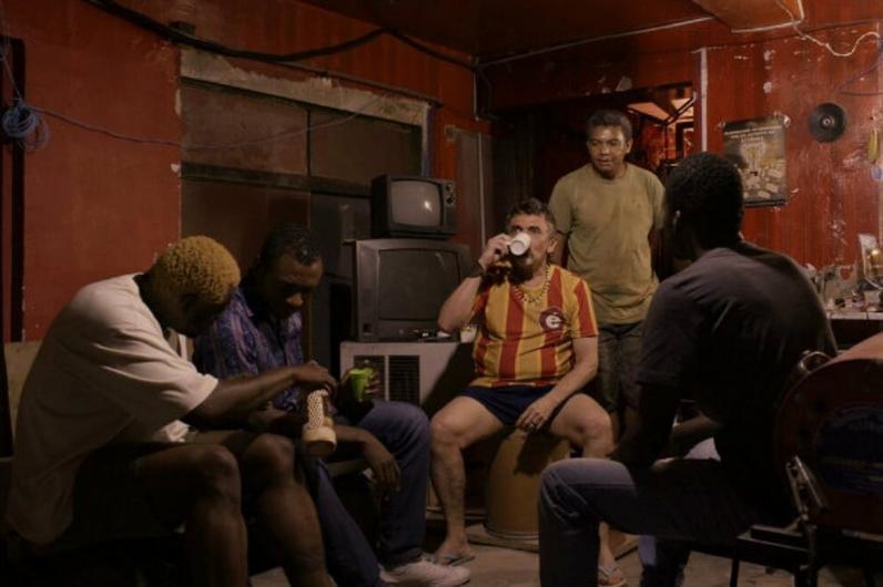 Imagem de uma cena do filme Era o Hotel Cambridge (2016). Quatro homens sentados e outro em pé, sendo um branco e os demais negros, encontram-se em uma sala pequena e apertada. Suas paredes são vermelhas e estão descascadas. O ambiente é pobre e mal iluminado. Ao fundo, perto de uma porta, duas televisões estão empilhadas sobre um móvel. 