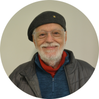 Foto de homem branco e idoso sorrindo. Ele tem olhos castanhos, cabelos curtos e grisalhos, barba grisalha, usa uma boina escura, óculos de armação retangular e transparente e blusas de gola alta vermelha e azul sob jaqueta cinza.