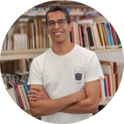 oto de um homem de braços cruzados sorrindo. Ele é pardo, tem olhos castanhos, cabelos curtos, lisos e levemente grisalhos, usa óculos com armação circular e camiseta branca com estampa no peito de um copo americano com café. Atrás dele, há uma estante vazada com livros.