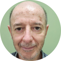 Foto de homem branco esboçando um sorriso. Ele tem os olhos castanhos, é calvo e tem barba e cabelos muito curtos grisalhos. Ele usa uma camisa xadrez azul e o fundo é verde claro. 