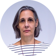 Foto de uma mulher branca de cabelos lisos e grisalhos, quase até os ombros. Ela usa colar e camiseta branca com linhas horizontais pretas. O fundo da foto é claro.