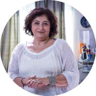 Foto de uma mulher branca de cabelos curtos avermelhados. Ela segura uma mão sobre a outra, usa um colar, pulseiras e blusa branca. A foto é em um ambiente interno, com cortinas verdes ao fundo, uma mesa pequena com vários utensílios, um espelho e relógio pendurados ao fundo.