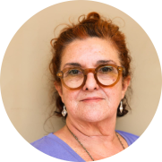 oto em moldura circular de uma mulher  com cabelos ondulados e ruivos, que estão presos. Ela usa óculos de armação redonda e marrom, veste blusa lilás e usa brincos prateados e um colar. O fundo é bege.